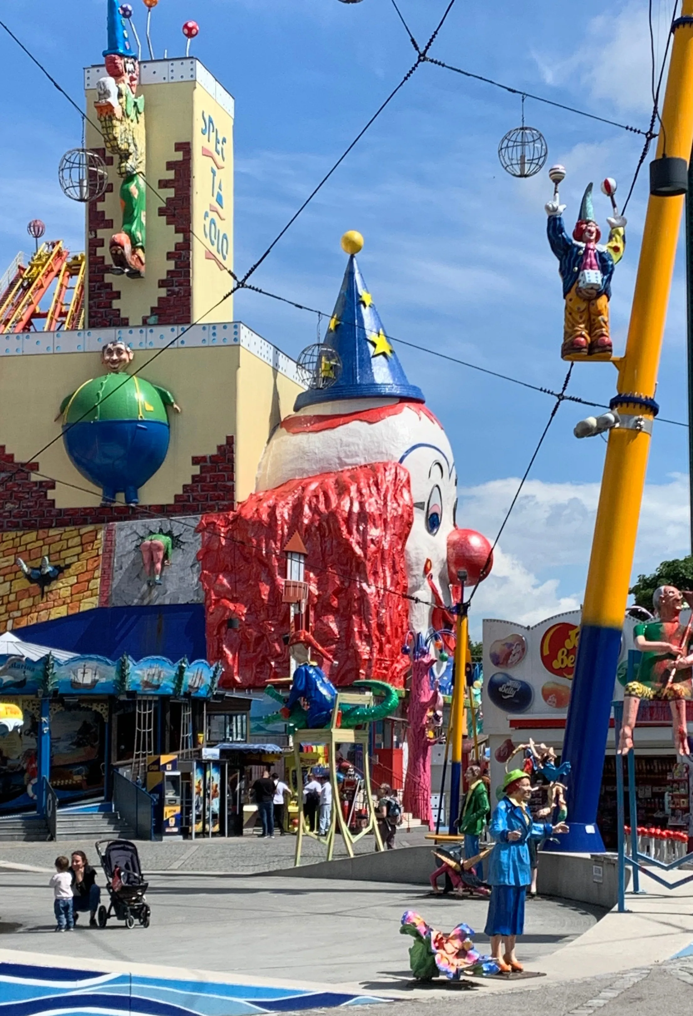 Ein farbenfroher Vergnügungspark mit übergroßen Clown-Statuen, einem Riesenrad und bunten Fahrgeschäften unter blauem Himmel. Menschen spazieren herum und genießen die festliche Atmosphäre.