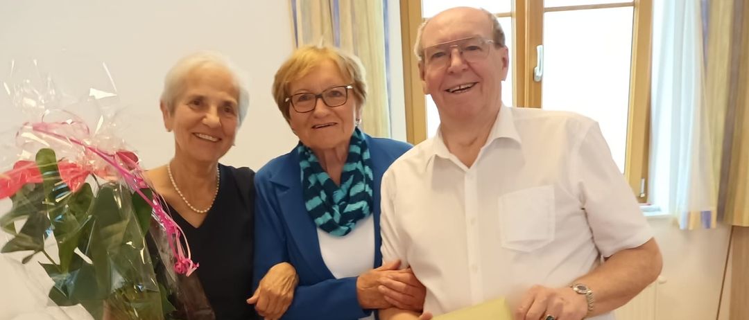 Drei Menschen stehen eng beieinander in einem Raum. Die Frau auf der linken Seite hält einen Blumenstrauß. Der Mann auf der rechten Seite hält einen gelben Umschlag.