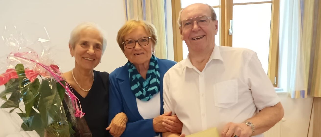 Drei Menschen stehen eng beieinander in einem Raum. Die Frau auf der linken Seite hält einen Blumenstrauß. Der Mann auf der rechten Seite hält einen gelben Umschlag.