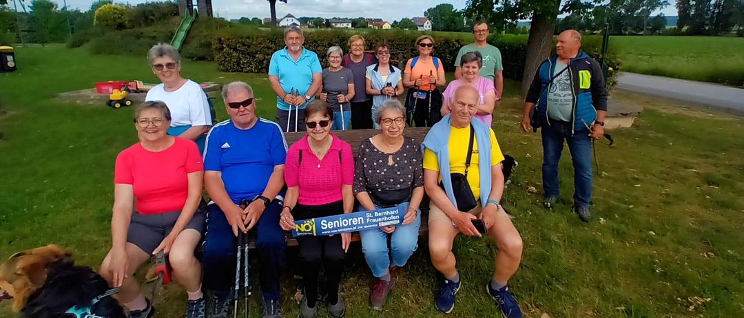 Eine Gruppe von Menschen, möglicherweise auf einer Wanderung, posiert für ein Foto auf einem Grasfeld mit einem Schild im Vordergrund. Sie tragen Freizeitkleidung und einige haben Wanderstöcke. Im Hintergrund steht ein Baum und ein Gebäude.