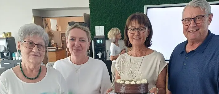 Vier ältere Personen versammeln sich lächelnd um einen Tisch. Eine Frau hält einen Geburtstagskuchen, während eine andere ein Schild mit der Aufschrift 'Senioren Horn' hält. Auf dem Tisch befinden sich eine Tasse, eine Untertasse und ein Papierbeutel. Im Hintergrund sind ein Kühlschrank und eine Tür zu sehen.