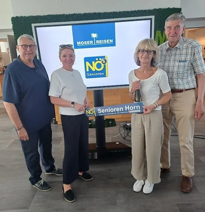 Vier Erwachsene stehen vor einem großen Monitor, der MOSER Reisen bewirbt, wobei zwei Frauen ein Schild und eine Flasche halten. Die Frauen lächeln.