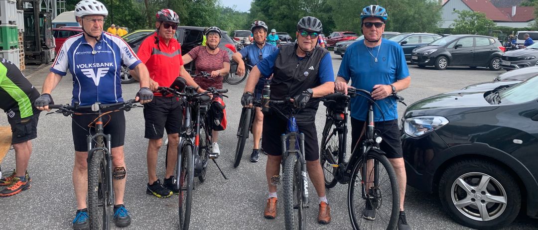 Eine Gruppe von Radfahrern posiert mit ihren Fahrrädern auf einem Parkplatz. Sie tragen Helme und Radbekleidung. Hinter ihnen sind einige Autos geparkt.