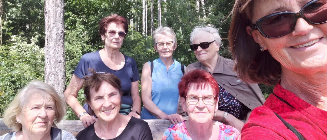 Eine Gruppe von sechs Frauen, einige mit Brille, lächeln für ein Foto im Wald. Sie stehen und sitzen auf einer Holzbank.