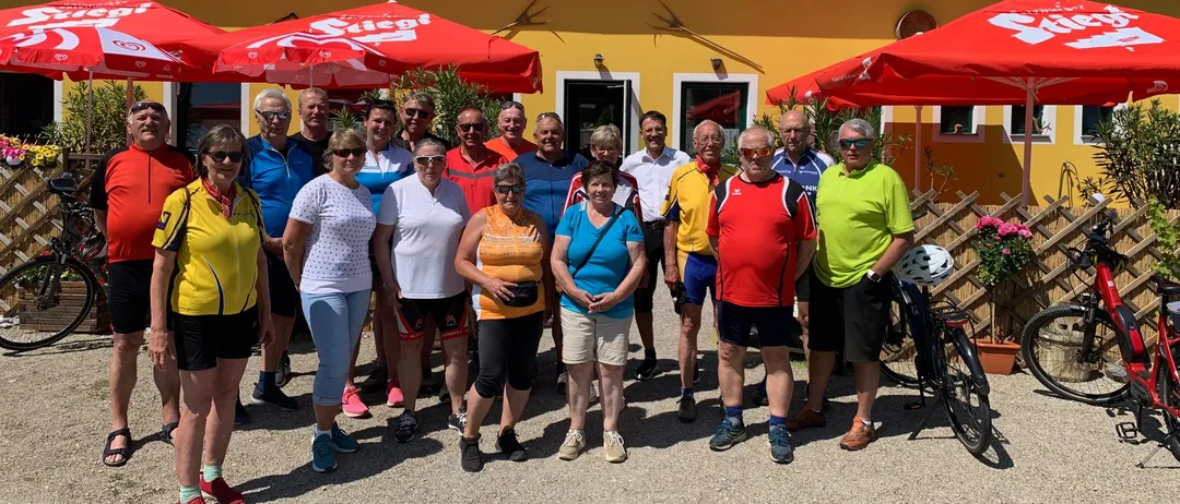 Eine Gruppe von Menschen in Fahrradbekleidung steht vor einem gelben Gebäude mit roten Dächern und Fenstern, unter roten Sonnenschirmen. Ein Fahrrad steht in der Nähe.