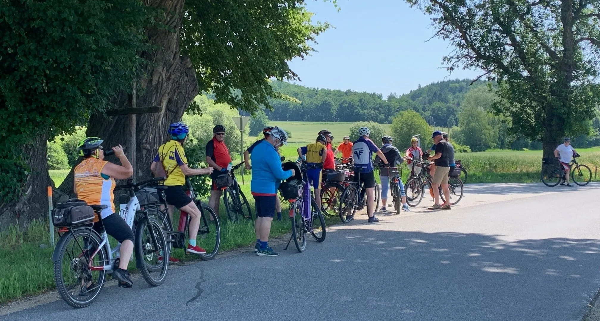 Eine Gruppe von Radfahrern steht am Straßenrand, alle tragen Helme und Radbekleidung. Einige haben Wasserflaschen dabei, einer trägt eine blaue Jacke. Im Hintergrund sind Bäume und ein grünes Feld zu sehen.