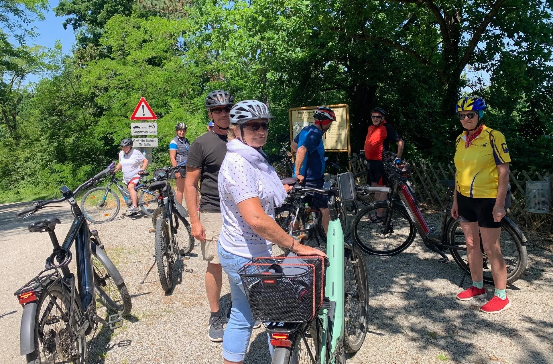 Eine Gruppe von Radfahrern mit Helmen und Fahrradausrüstung versammelt sich in einem Waldgebiet. Bäume und ein Schild sind im Hintergrund sichtbar. Sie stehen mit ihren Fahrrädern, wahrscheinlich machen sie eine Pause.