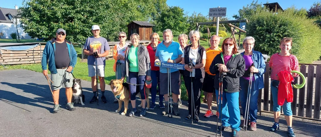 Eine Gruppe von Seniorenwanderern posiert an einem sonnigen Tag für ein Foto. Sie sind für Outdoor-Aktivitäten gekleidet und werden von Hunden begleitet. Ein Schild im Hintergrund lautet 'Ellern hallen für ihre Kinder.'