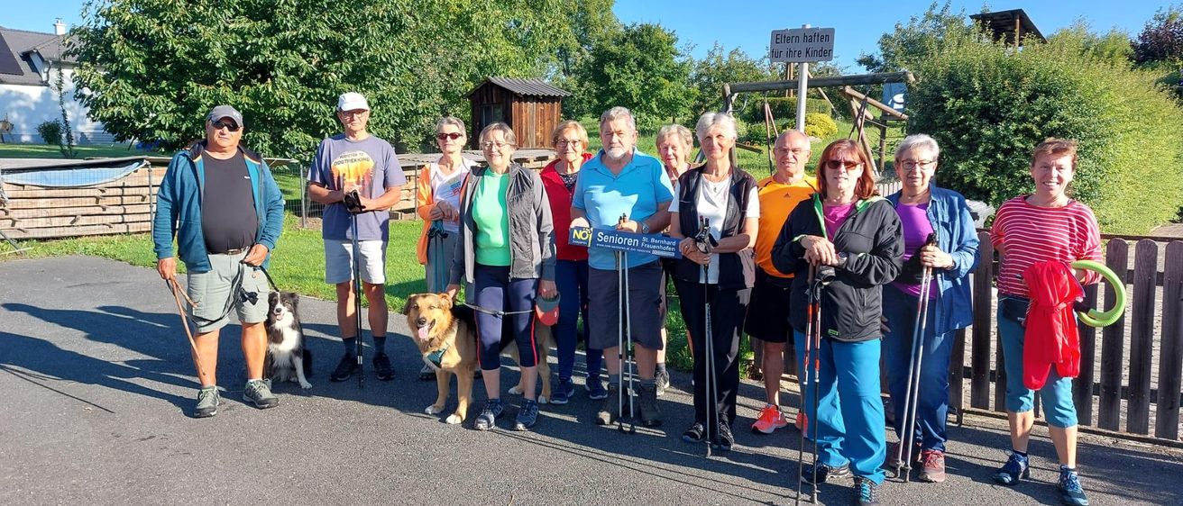 Eine Gruppe von Seniorenwanderern posiert an einem sonnigen Tag für ein Foto. Sie sind für Outdoor-Aktivitäten gekleidet und werden von Hunden begleitet. Ein Schild im Hintergrund lautet 'Ellern hallen für ihre Kinder.'
