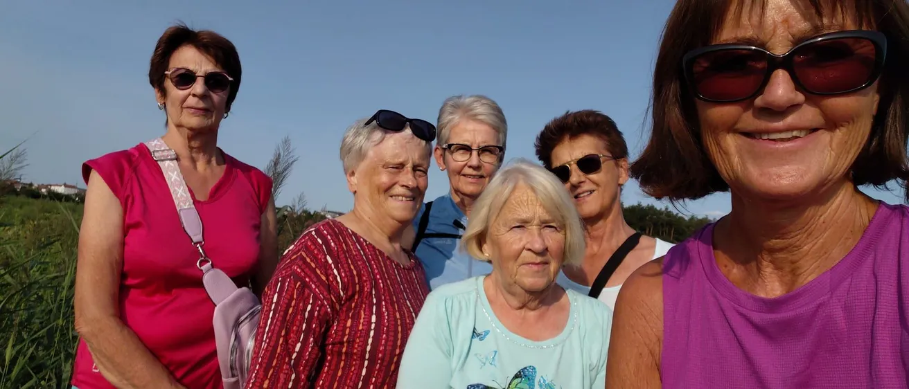 Eine Gruppe lächelnder älterer Frauen, alle mit Sonnenbrille, steht draußen. Sie sind in Freizeitkleidung gekleidet und scheinen an einem sonnigen Tag zu sein.