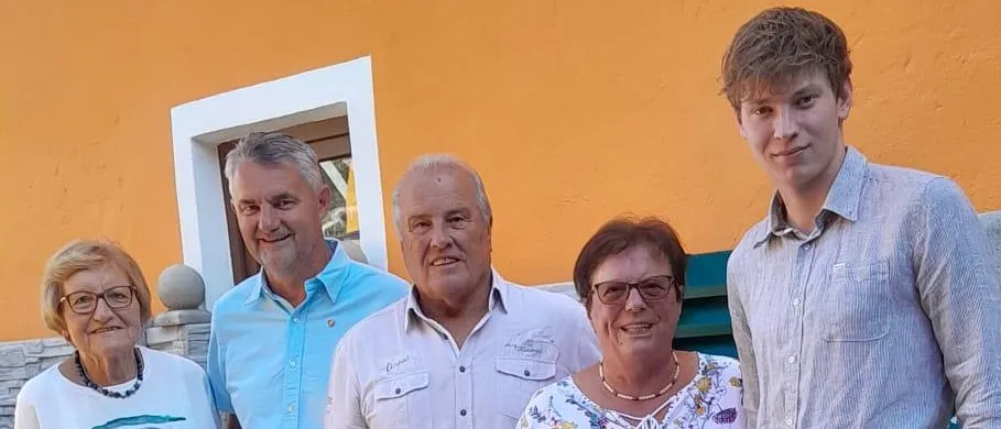 Fünf Personen posieren lächelnd für ein Foto auf einer Ziegelterrasse. Eine Frau hält einen Blumenstrauß. Hinter ihnen sind Topfpflanzen und eine gelbe Wand zu sehen.