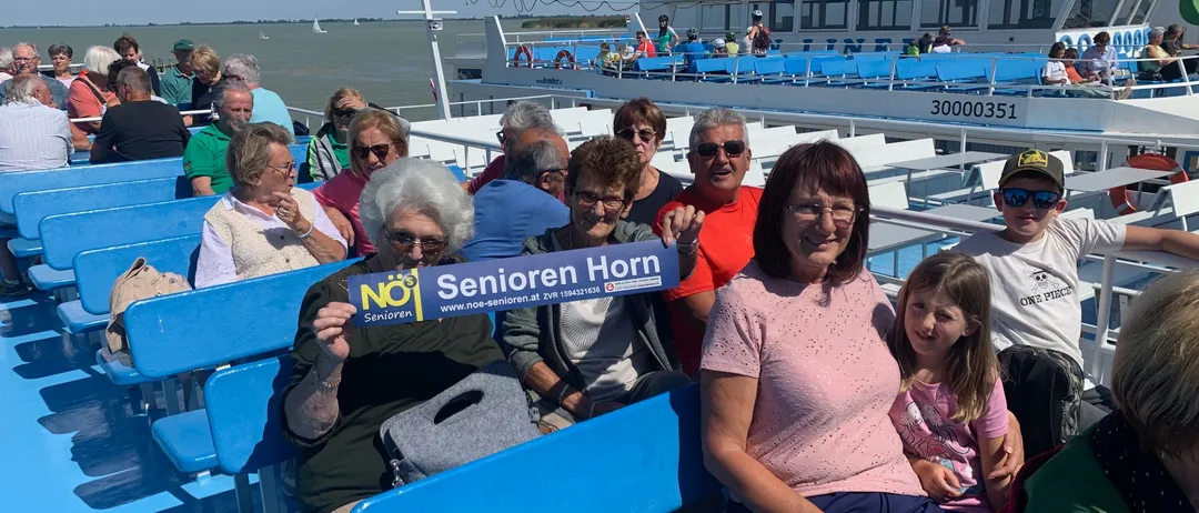 Gruppe älterer Personen auf einem Boot, einige halten ein blaues Schild mit der Aufschrift 'Senioren Horn', sitzen auf blauen Sitzen mit einem Gebäude im Hintergrund.