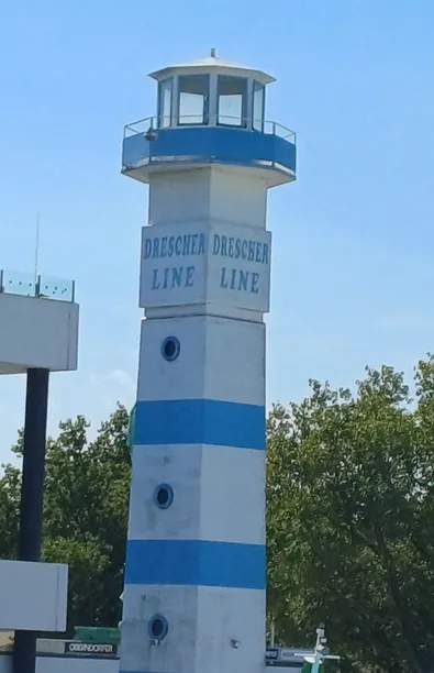 Eine Leuchtturm-ähnliche Struktur mit blauen und weißen Streifen trägt zweimal den Namen 'Dreschner' und zweimal 'Line'. Sie ist von Bäumen umgeben.