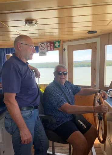 Zwei ältere Männer sind auf einem Boot, einer steuert und der andere steht neben ihm. Das Boot fährt auf einem Gewässer mit Blick auf das Land.