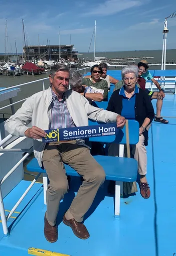Ein älteres Paar und drei weitere Personen sitzen auf dem Deck eines Bootes. Sie alle lächeln. Der Mann hält ein blaues Schild.