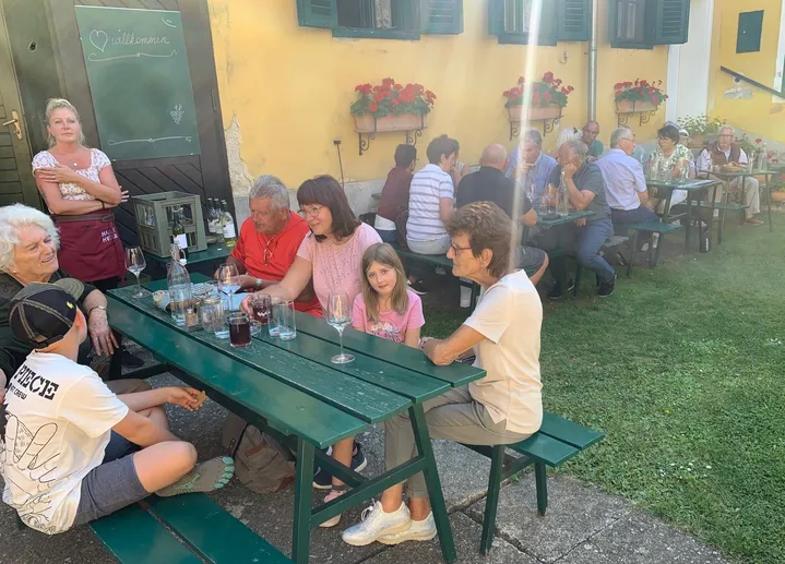 Ein Outdoor-Essbereich mit einer Gruppe älterer Menschen, die an grünen Picknicktischen sitzen, einem Tafelschild im Hintergrund und roten Blumen in Pflanzgefäßen an der Wand.