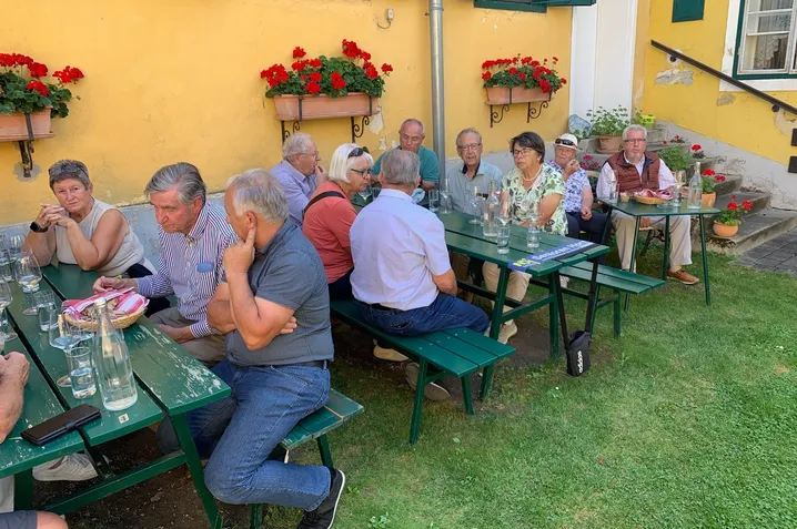 Eine Gruppe älterer Menschen sitzt um grüne Tische in einem Außengarten. Auf den Tischen stehen Topfpflanzen und Gläser.