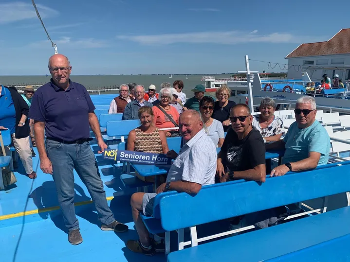 Eine Gruppe älterer Menschen ist auf einer blauen Fähre, lächelt und schaut in die Kamera. Der Mann in der Mitte hält ein blaues Schild mit weißer Schrift.