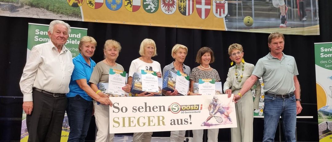 Eine Gruppe von Personen hält Urkunden in der Hand und posiert für ein Foto auf der Bühne. Der Banner hinter ihnen lautet 'So sehen SIEGER aus!'. Sie tragen alle Turnschuhe.