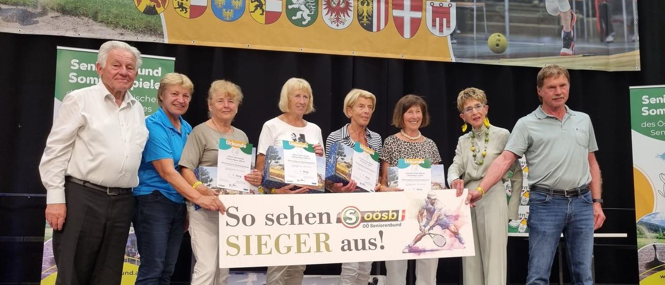 Eine Gruppe von Personen hält Urkunden in der Hand und posiert für ein Foto auf der Bühne. Der Banner hinter ihnen lautet 'So sehen SIEGER aus!'. Sie tragen alle Turnschuhe.