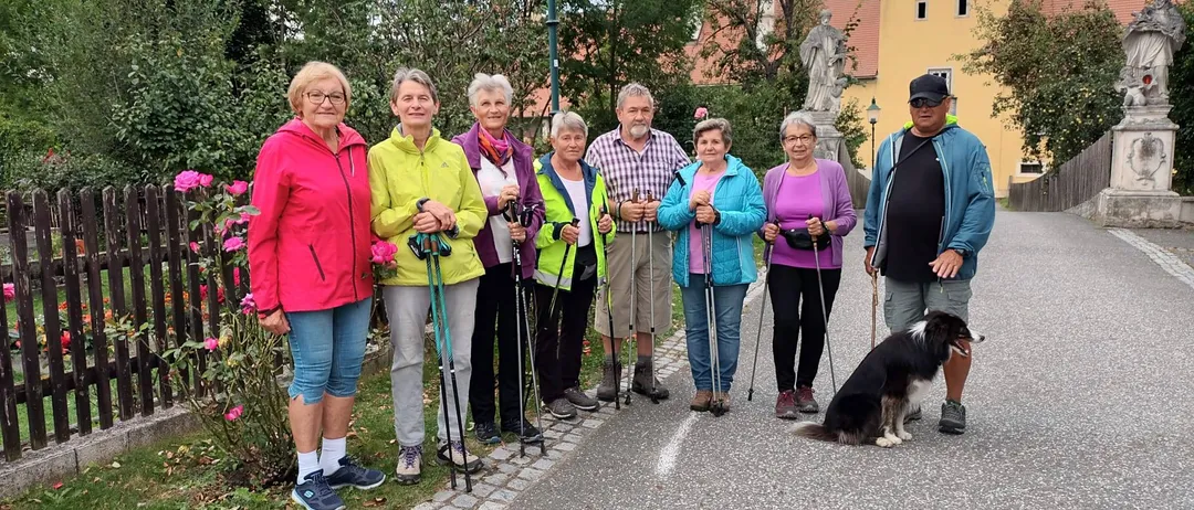 Eine Gruppe von Menschen steht auf einer Straße mit einem Hund und hält alle Wanderstöcke. Dahinter sind eine Statue und ein Gebäude mit rotem Dach zu sehen.