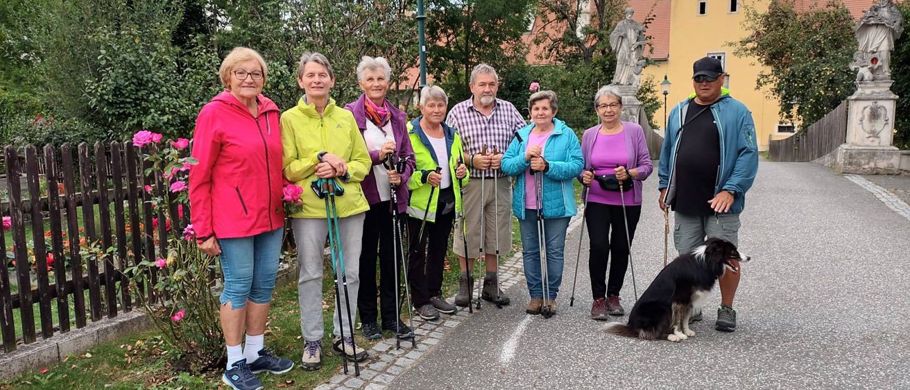 Eine Gruppe von Menschen steht auf einer Straße mit einem Hund und hält alle Wanderstöcke. Dahinter sind eine Statue und ein Gebäude mit rotem Dach zu sehen.