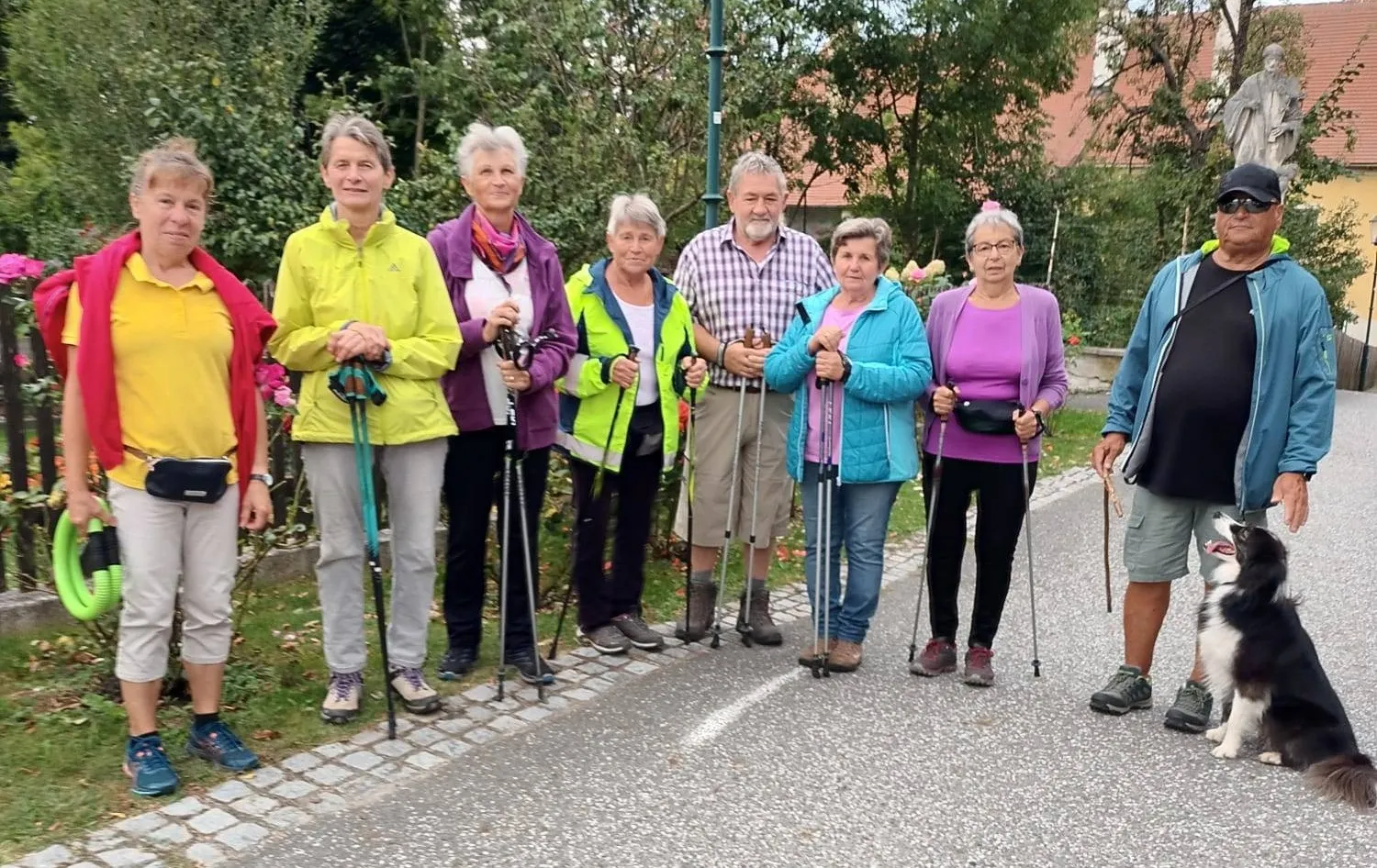 Eine Gruppe älterer Personen steht auf einer Straße, hält Wanderstöcke in der Hand und ist in verschiedene Jacken gekleidet. Sie nehmen wahrscheinlich an einer Wanderveranstaltung teil. Bäume und ein Gebäude sind im Hintergrund zu sehen.