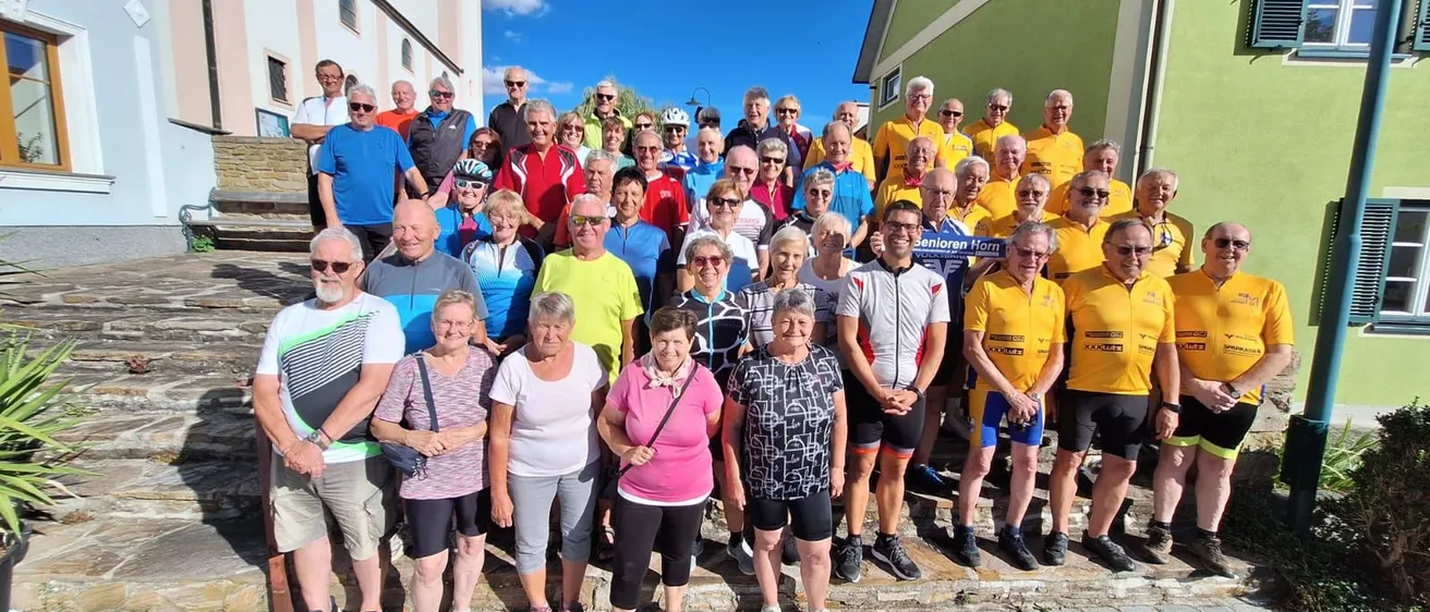 Eine Gruppe von Menschen, einige mit Sonnenbrillen und Fahrradtrikots, stehen auf Kopfsteinpflaster vor einem Gebäude unter einem teilweise bewölkten Himmel.