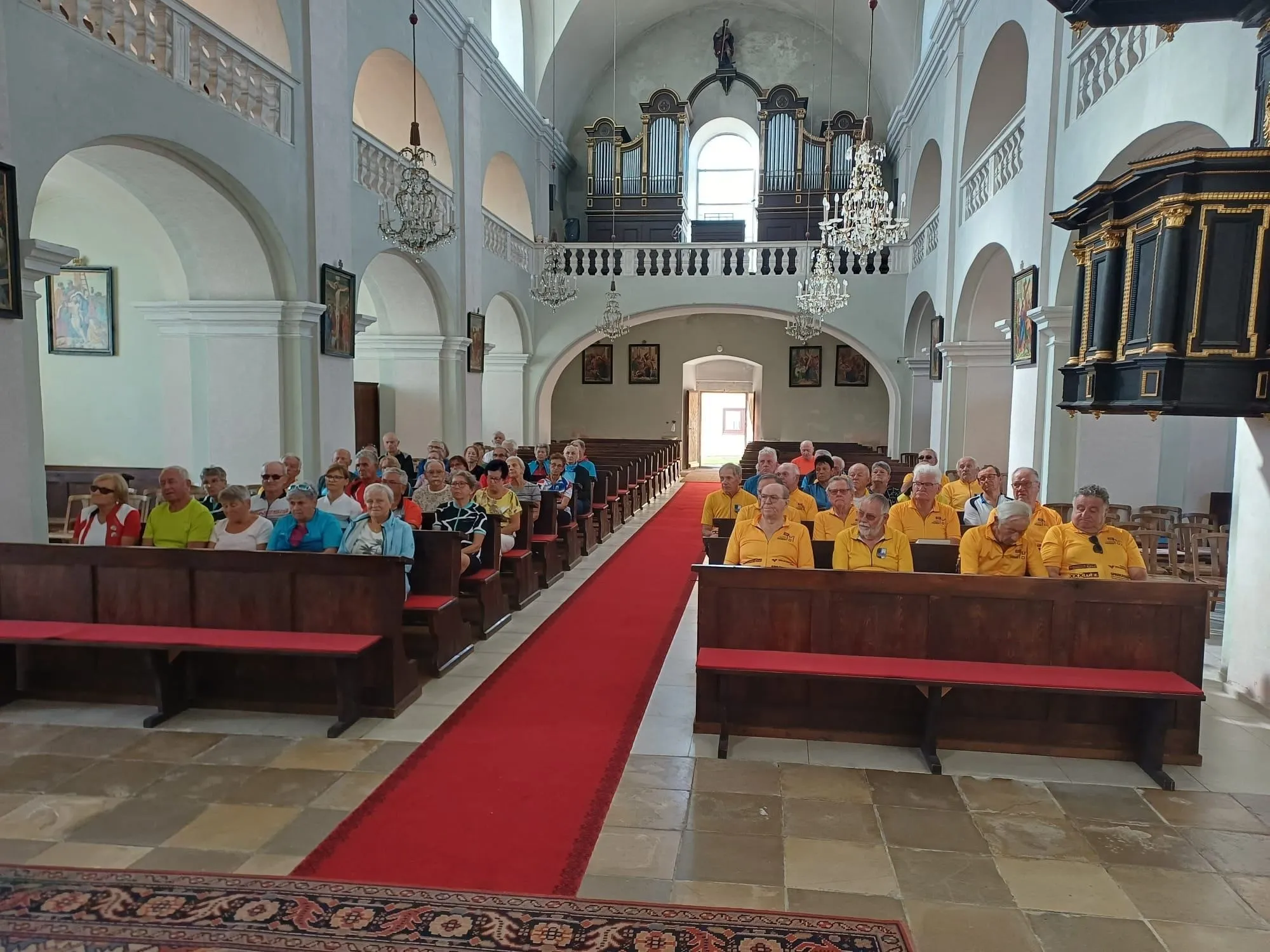 Eine Gruppe von Menschen in gelben Westen sitzt in Kirchenbänken, mit einem roten Teppich zum Altar. Kronleuchter hängen von der Decke, und mehrere Gemälde schmücken die Wände.