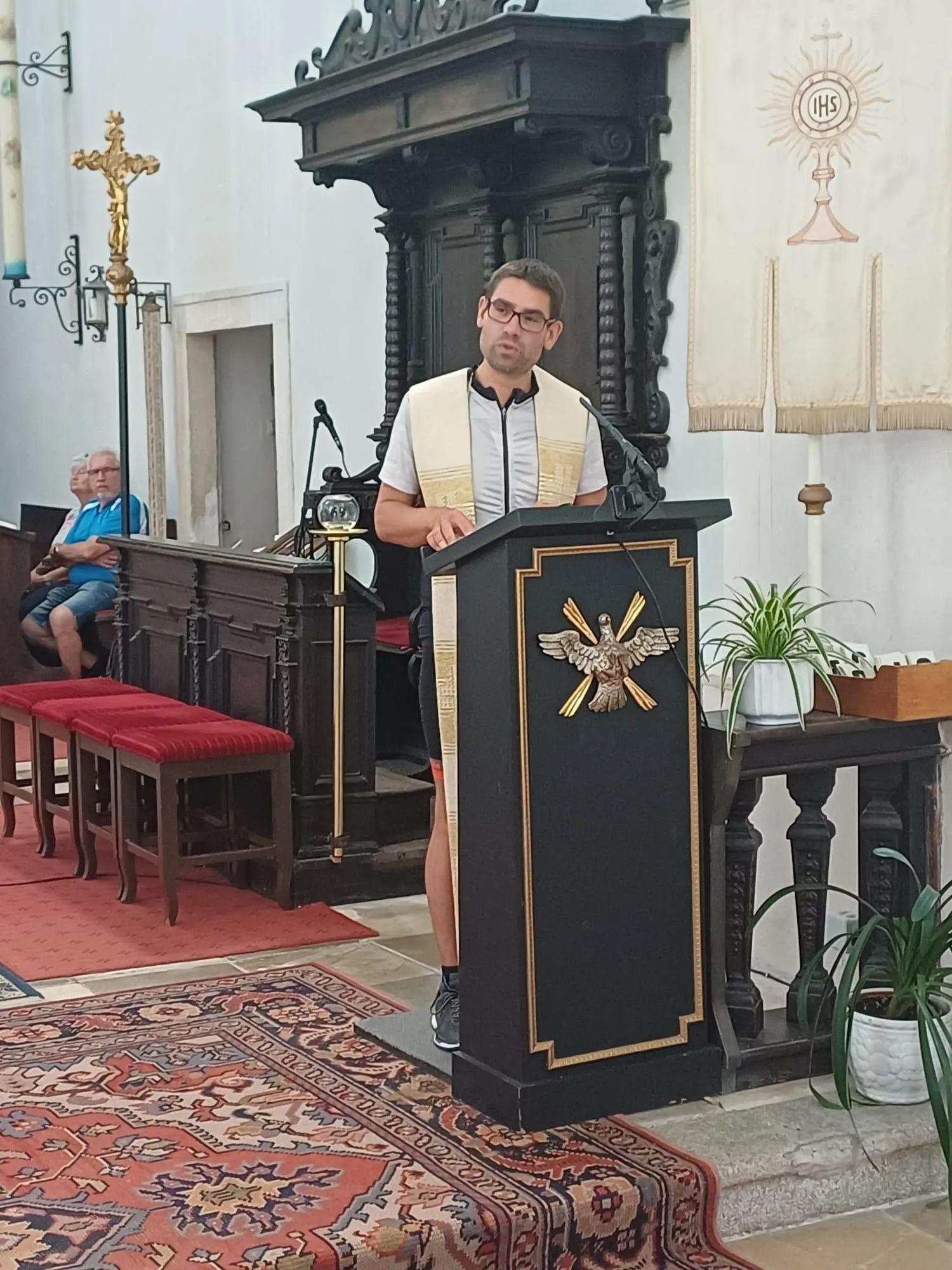 Ein Mann steht in einer Kirche an einem Pult und trägt eine Brille und eine Stola. Hinter ihm sitzen zwei Personen auf einer Bank, und eine Lampe hängt von der Decke. Auf beiden Seiten des Pults befinden sich Holzbänke und eine Pflanze in einem Topf.