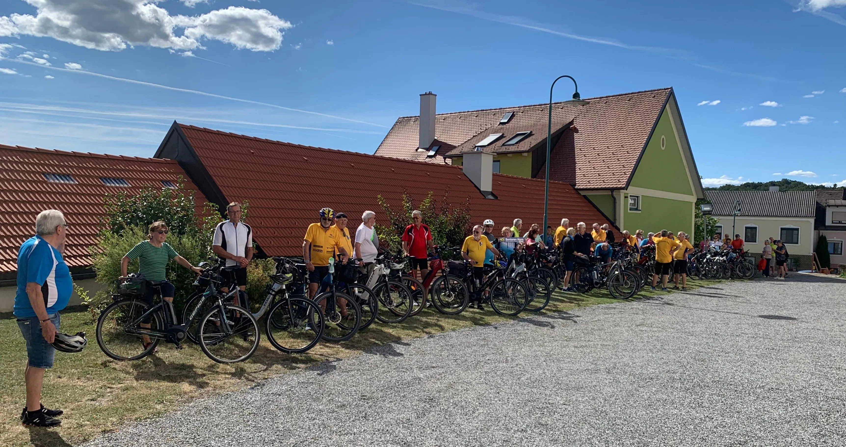 Eine Gruppe von Radfahrern steht vor einem Haus mit rotem Dach unter einem klaren blauen Himmel mit ein paar Wolken. Sie tragen alle Helme und Fahrradausrüstung.