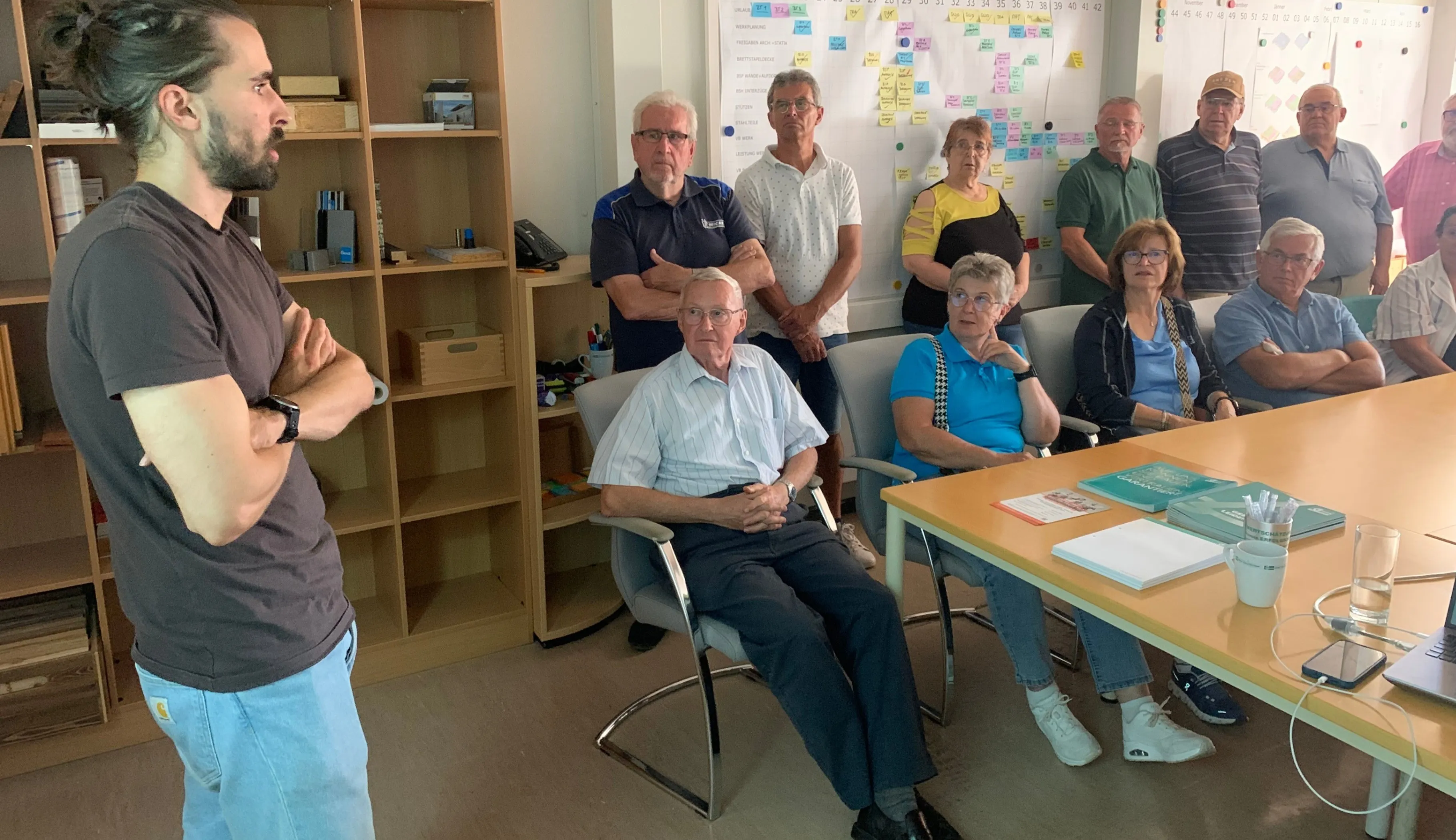 Eine Gruppe von Menschen in einem Besprechungsraum mit einer Tafel, Büchern und einem Kalender. Ein Mann sitzt im Vordergrund, und andere stehen hinter ihm.