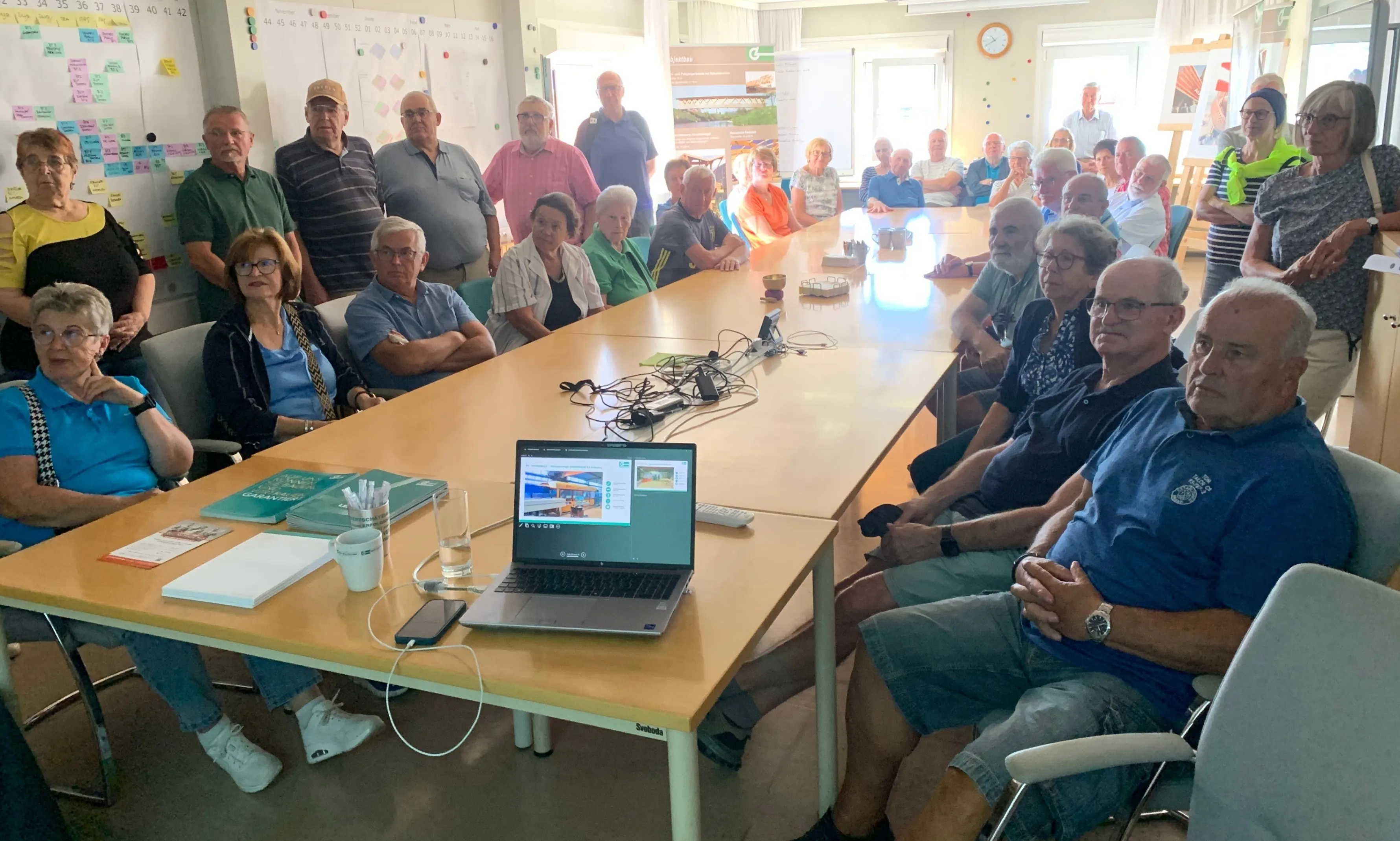Eine Gruppe von Menschen sitzt um einen Tisch mit einem Laptop. Einige tragen Brillen, Uhren und Turnschuhe. Eine Tafel hängt an der Wand.