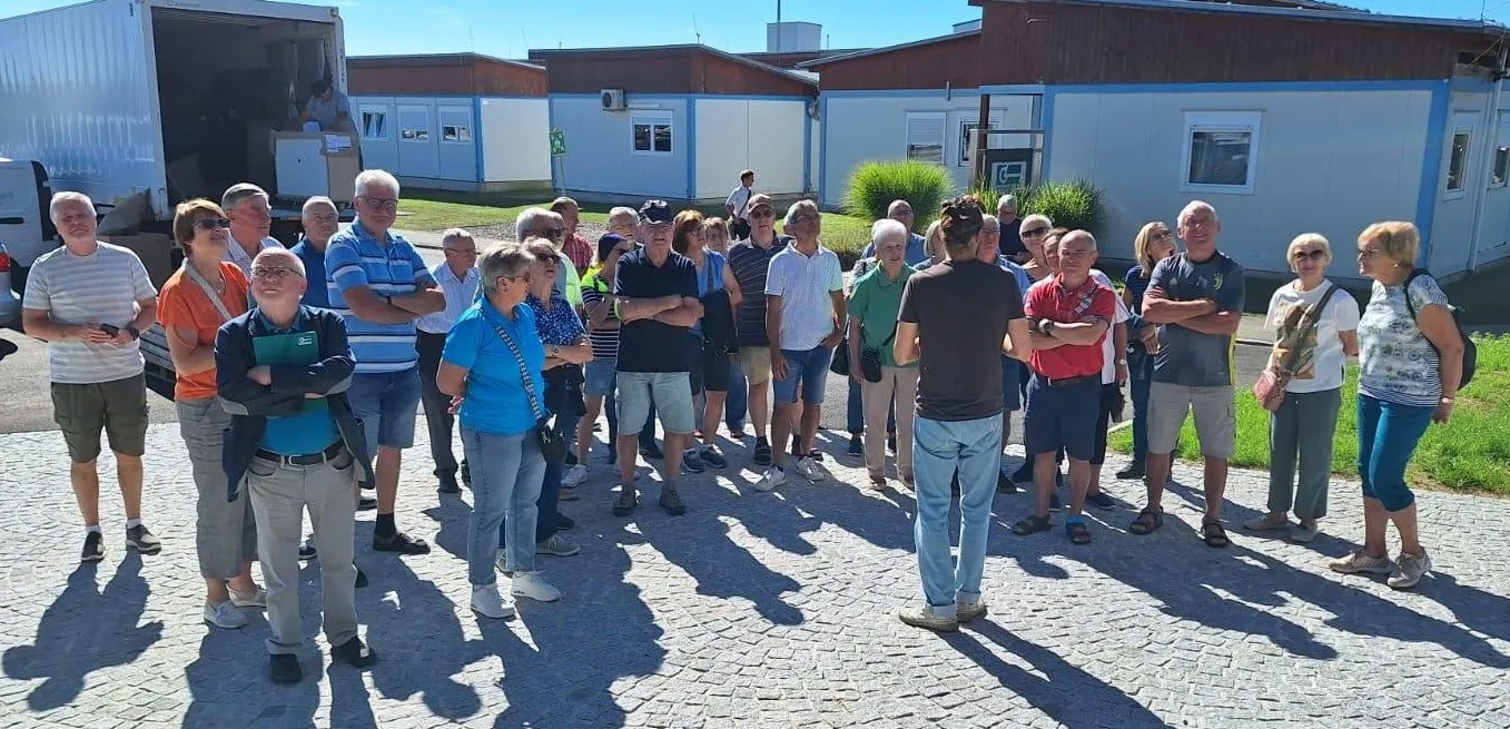 Eine Gruppe von Menschen steht auf einem Ziegelweg vor einem Gebäude. Einige tragen Brillen, Hüte und Turnschuhe. Ein Mann mit verschränkten Armen spricht sie an.