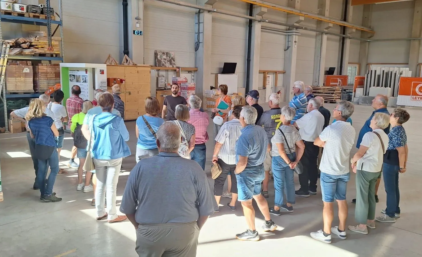 Eine Gruppe von Menschen steht in einem Raum, möglicherweise in einer Diskussion. Einige tragen Brillen und einige haben Handtaschen dabei. Der Raum hat eine weiße Wand und Decke mit einem Rohr. Ein Mann steht vor einem Holzschrank mit einem Monitor obenauf.