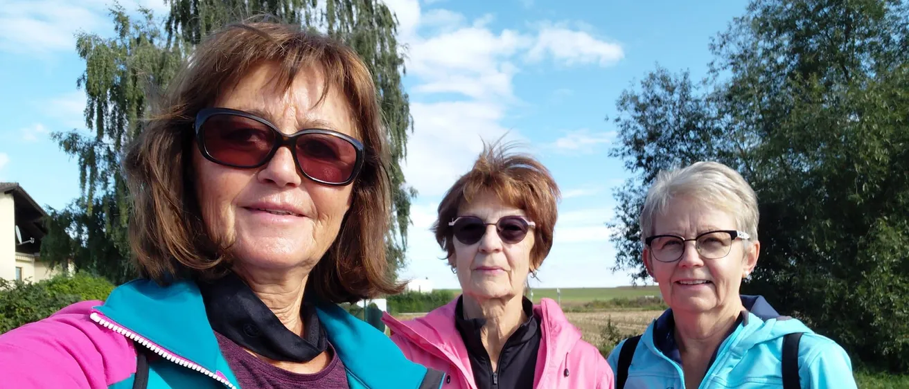 Drei Frauen in Jacken und mit Sonnenbrillen stehen auf einem Feld mit Bäumen und blauem Himmel im Hintergrund.