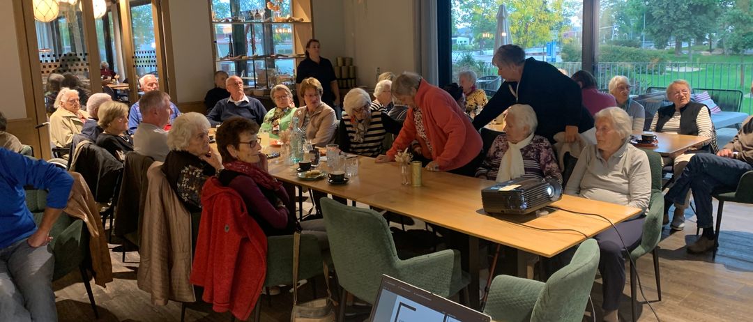 Eine Gruppe älterer Menschen ist in einem Restaurant versammelt und sitzt um einen langen Tisch mit einem Projektor herum. Sie unterhalten sich, während sie auf grünen Stühlen sitzen. Das Restaurant hat große Fenster, die Sonnenlicht hereinlassen.