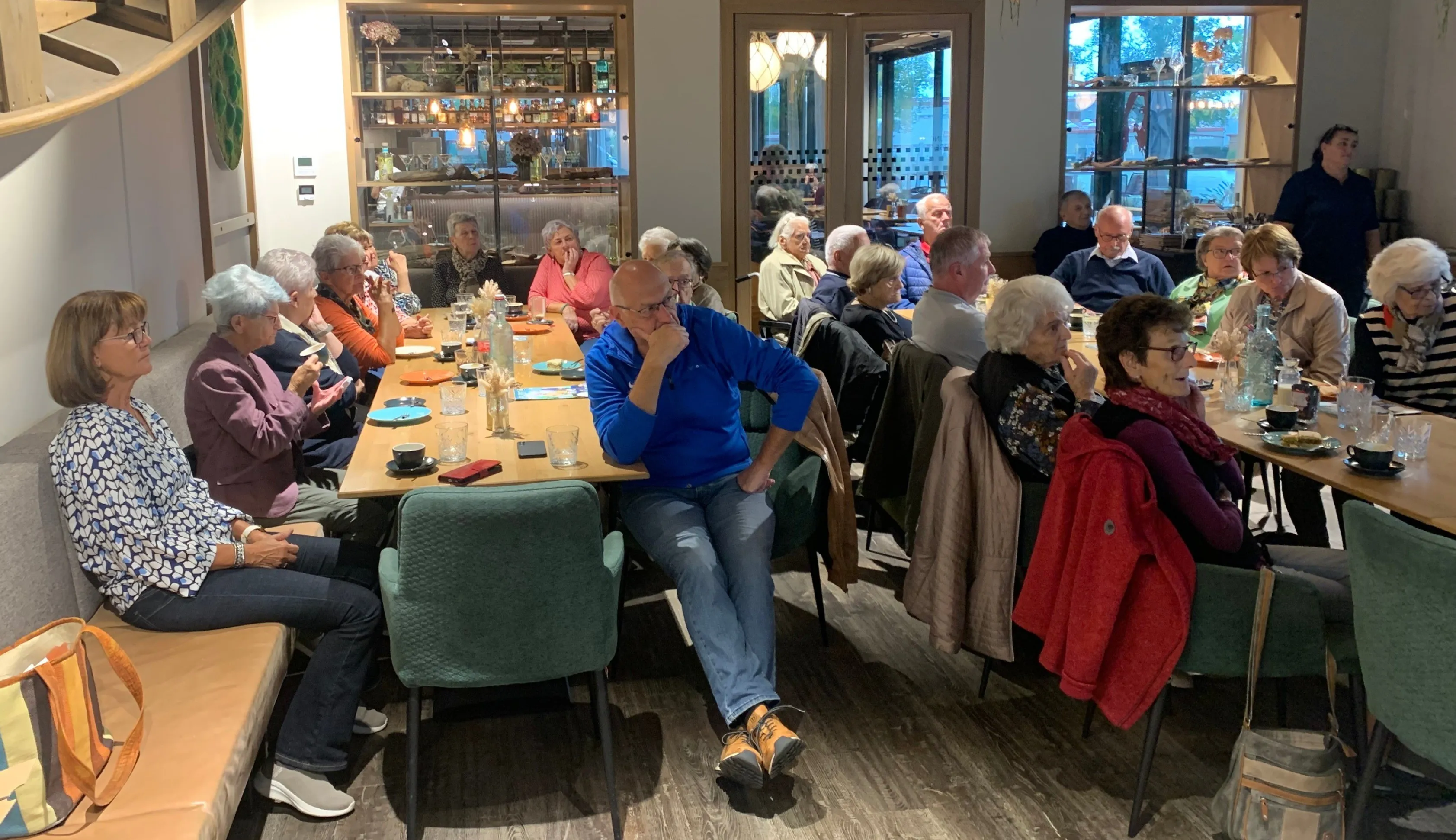 Eine Gruppe älterer Menschen sitzt um einen Tisch in einem Restaurant, einige mit den Händen am Kinn. Der Tisch ist mit Tellern, Gläsern und Besteck gedeckt. Ein Mann sitzt am Ende des Tisches mit den Händen am Kinn.