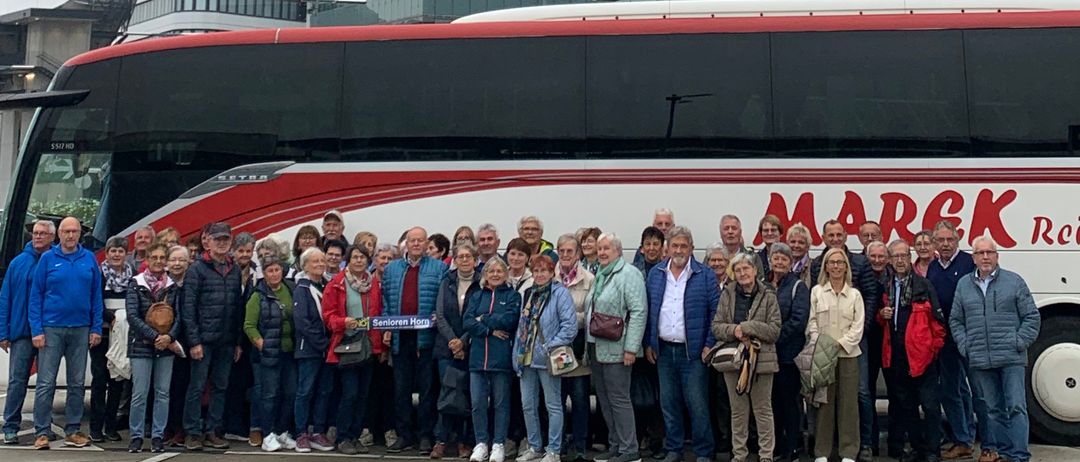 Eine Gruppe von Menschen in Winterkleidung steht vor einem modernen Gebäude und einem Bus. Der Bus hat das Wort 'MARCK' darauf. Dahinter befindet sich ein hoher Turm und andere Gebäude.
