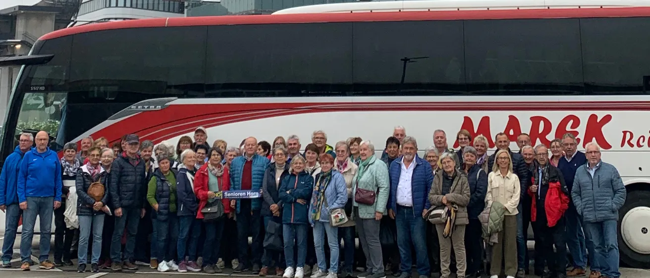 Eine Gruppe von Menschen in Winterkleidung steht vor einem modernen Gebäude und einem Bus. Der Bus hat das Wort 'MARCK' darauf. Dahinter befindet sich ein hoher Turm und andere Gebäude.