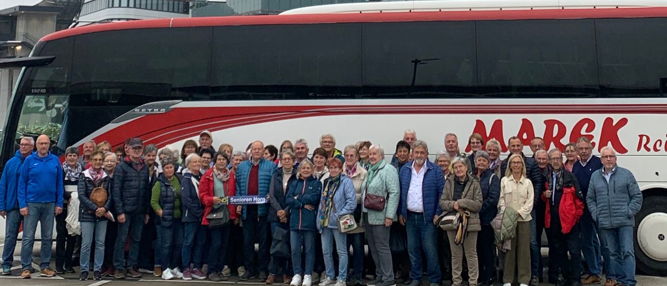 Eine Gruppe von Menschen in Winterkleidung steht vor einem modernen Gebäude und einem Bus. Der Bus hat das Wort 'MARCK' darauf. Dahinter befindet sich ein hoher Turm und andere Gebäude.