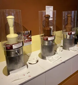 Drei Schokoladenbrunnen sind auf einer weißen Arbeitsplatte ausgestellt. Jeder Brunnen ist in einer Glasvitrine untergebracht und zeigt eine Schokoladenskulptur. Der linke ist weiß, die anderen sind dunkelbraun.