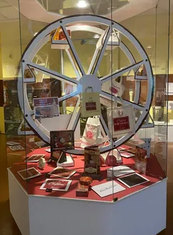 Eine Vitrine zeigt ein großes, dekoratives Rad, umgeben von Gegenständen wie Taschen und Kisten. Das Rad hat einen roten Rand und zeigt eine Vielzahl von Artikeln, darunter Bücher und Werbematerialien.