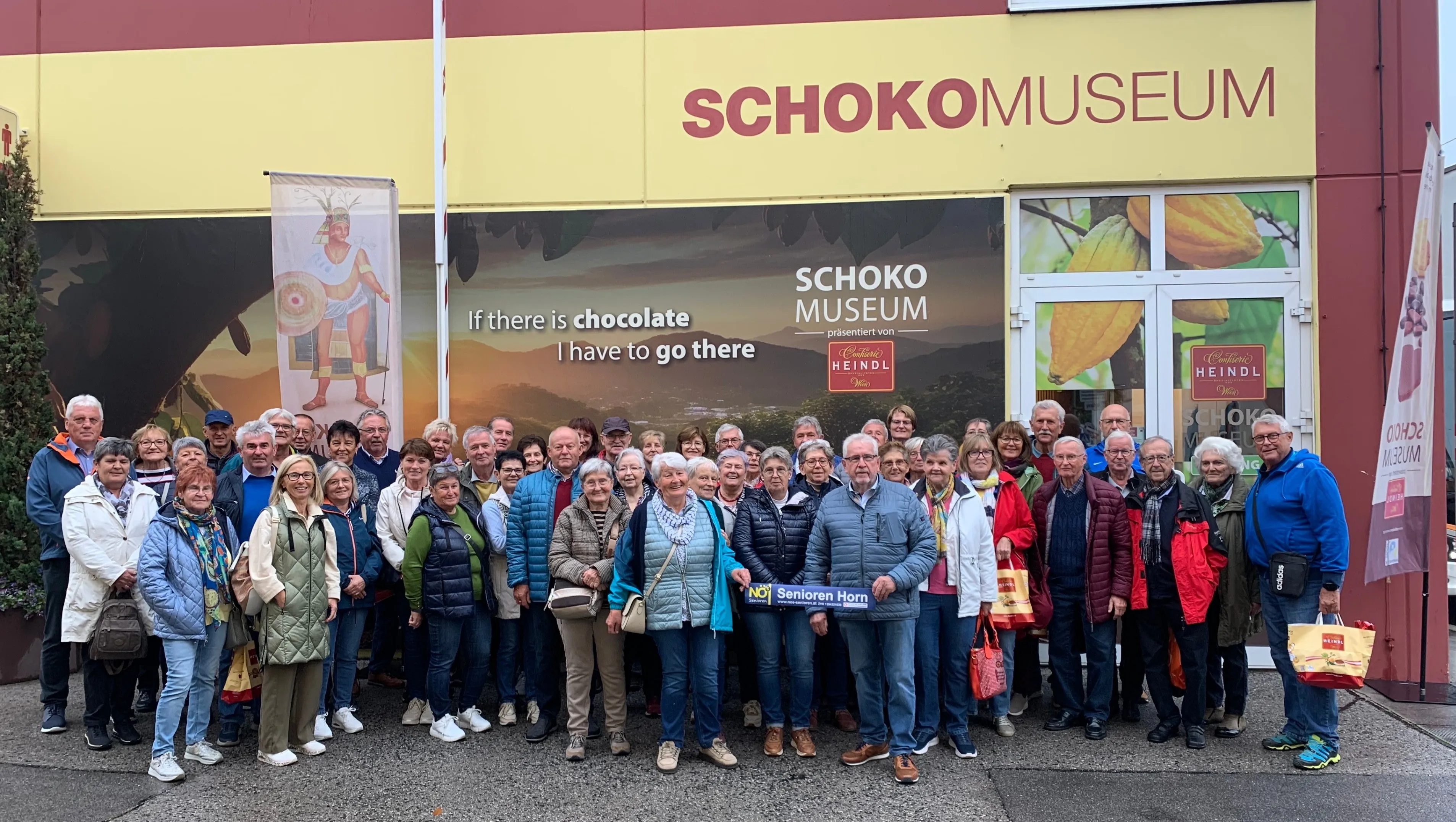 Eine Gruppe von Menschen steht vor einem Gebäude mit einem Schild, das 'Schoko Museum' und 'Wenn es Schokolade gibt, muss ich dorthin' besagt. Sie alle lächeln und scheinen für ein Foto zu posieren.