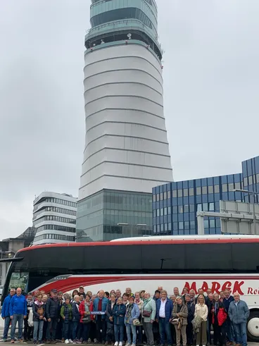 Eine Gruppe von Menschen steht vor einem rot-weißen Bus, lächelt und posiert für ein Foto. Dahinter befindet sich ein hoher weißer Turm und mehrere Gebäude.