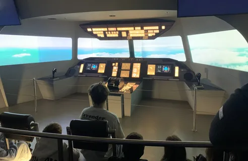 Eine Gruppe von Kindern sitzt in einem Flugsimulator. Die Person vorne trägt ein Lavendel-T-Shirt. Das Cockpit ist vollständig mit Monitoren und Bedienfeldern ausgestattet. Draußen ist der Himmel durch die Fenster zu sehen.