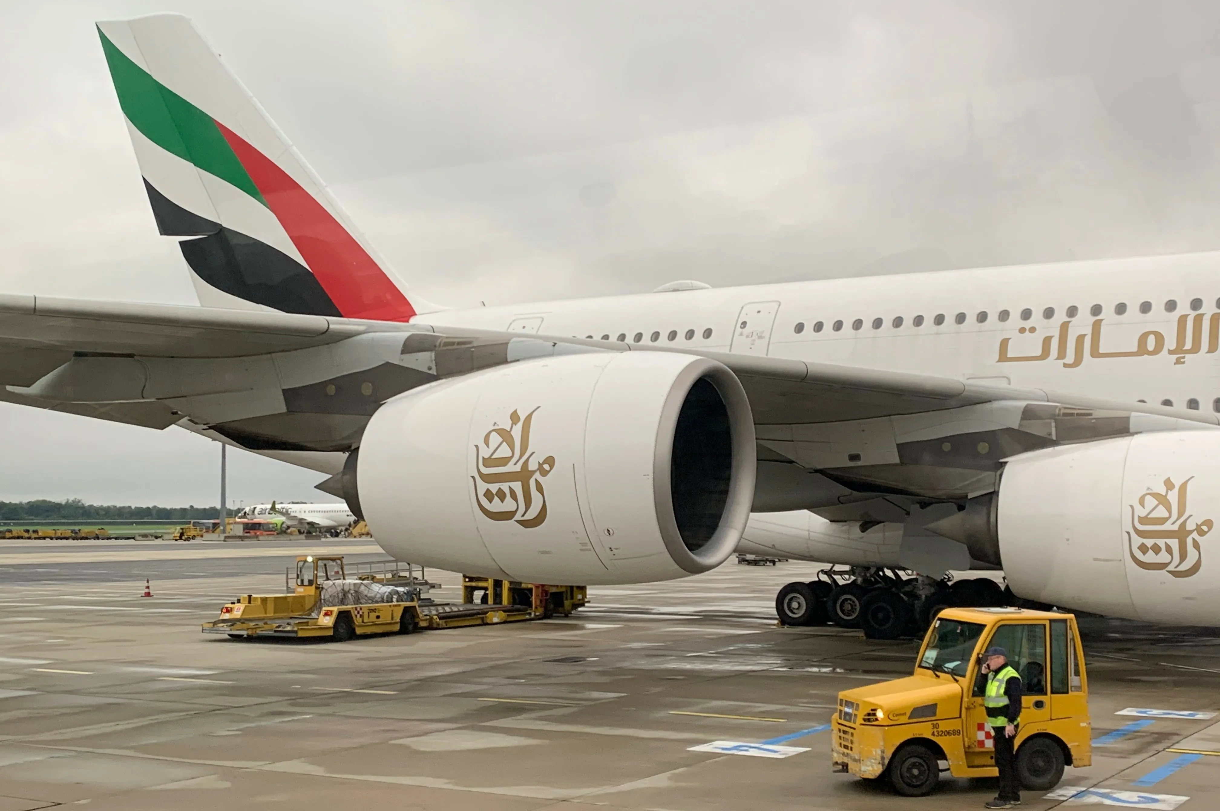 Ein Emirates-Flugzeug steht mit freiliegenden Triebwerken auf dem Vorfeld. Ein gelber Gepäckwagen bewegt sich in der Nähe.