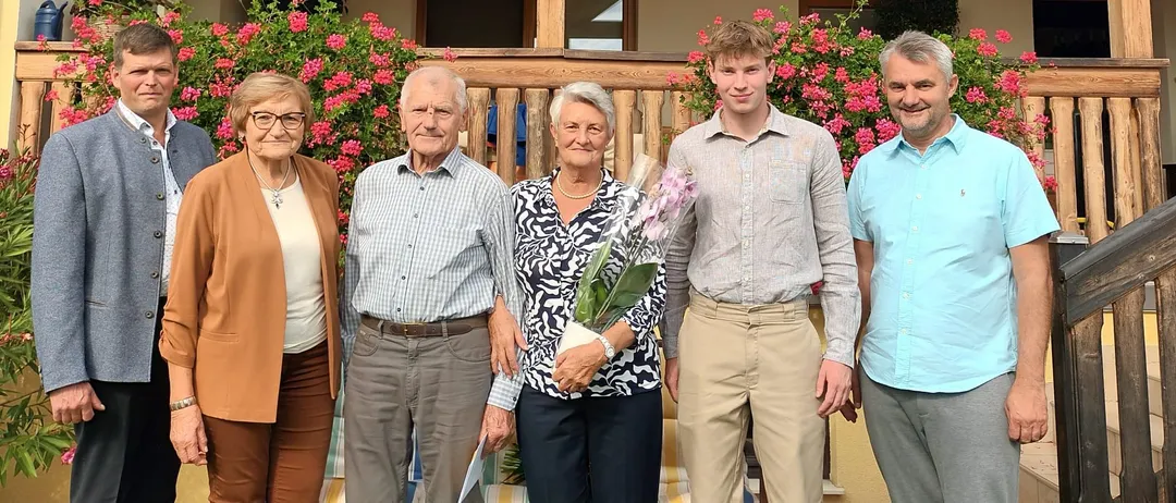 Eine Gruppe von sechs Personen steht vor einem Haus mit einer Veranda. Sie lächeln für ein Foto. Das Haus hat ein Holzdesign und ein Glasfenster.