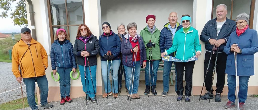 Eine Gruppe älterer Menschen steht in einer Reihe mit Wanderstöcken, in Winterkleidung. Sie lächeln und scheinen für ein Foto zu posieren. Hinter ihnen steht ein Gebäude mit Glasfenstern.
