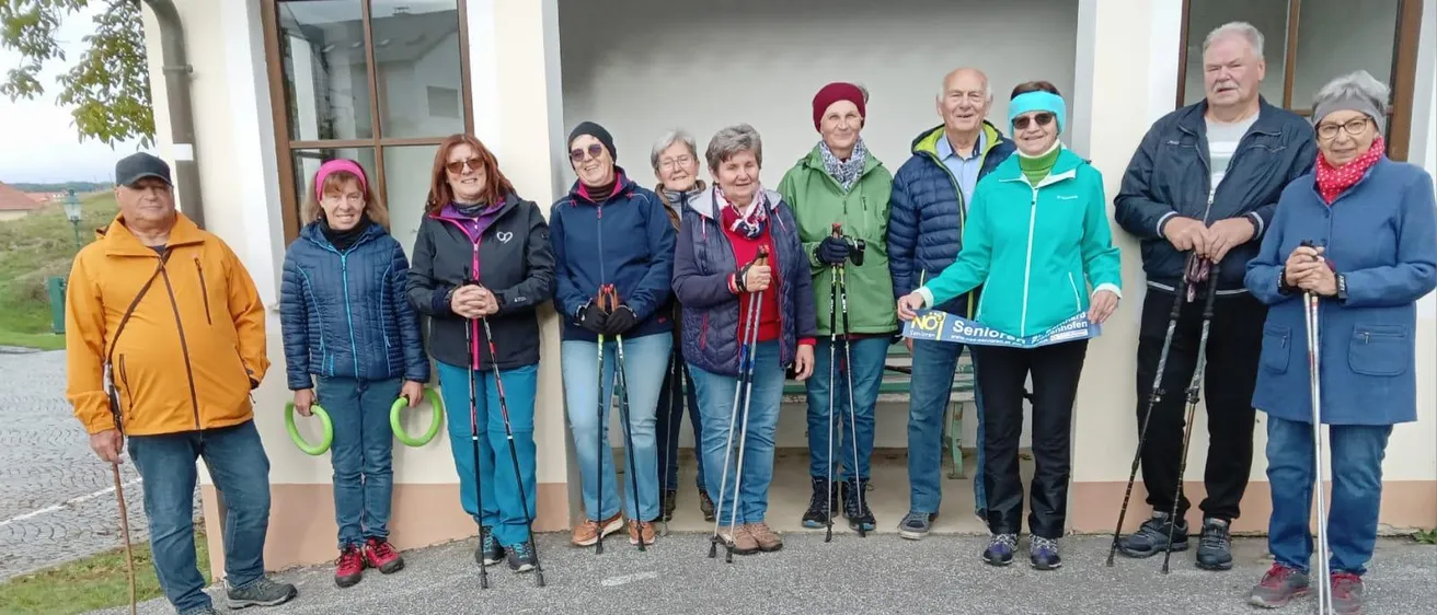 Eine Gruppe älterer Menschen steht in einer Reihe mit Wanderstöcken, in Winterkleidung. Sie lächeln und scheinen für ein Foto zu posieren. Hinter ihnen steht ein Gebäude mit Glasfenstern.